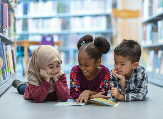 Three (3) Kids Who Are Friends Are Laying On The Floor In Between Bookshelves In The Library. They Are Reading A Book Together. They Are All Smiling And Enjoying Their Afternoon.