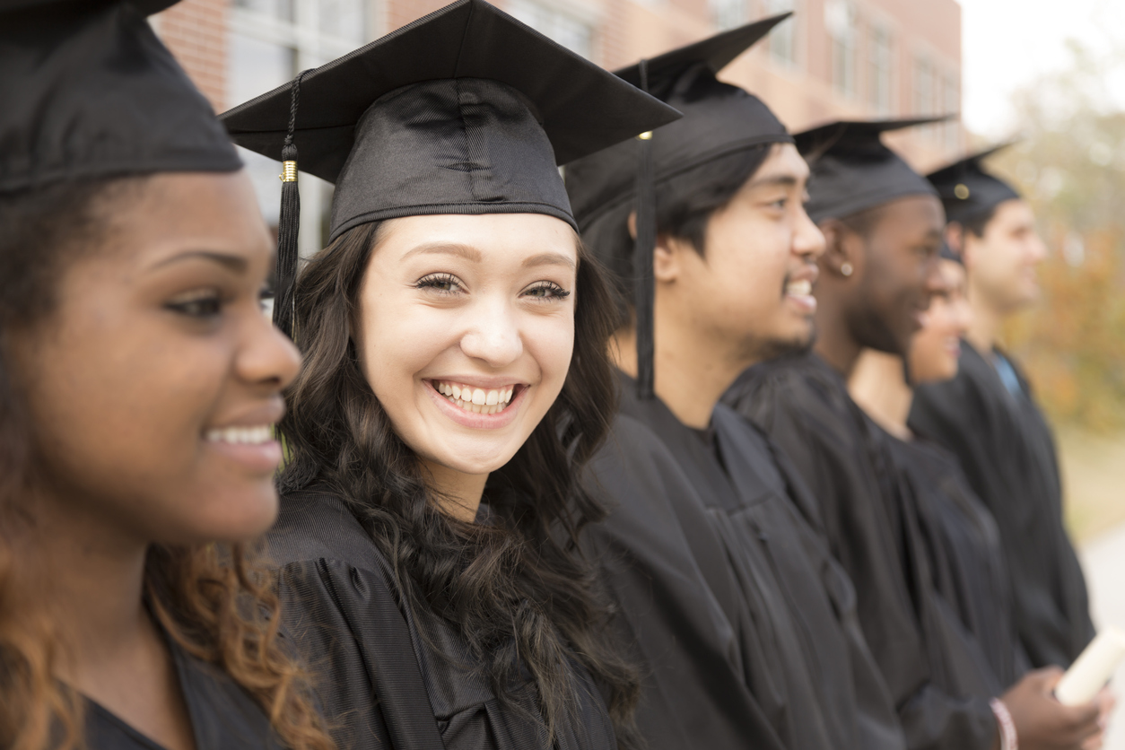 Six multi-ethnic friend graduates excitedly wait for their name to be called during graduation ceremony. Mixed-race girl looks back at camera. School building background.